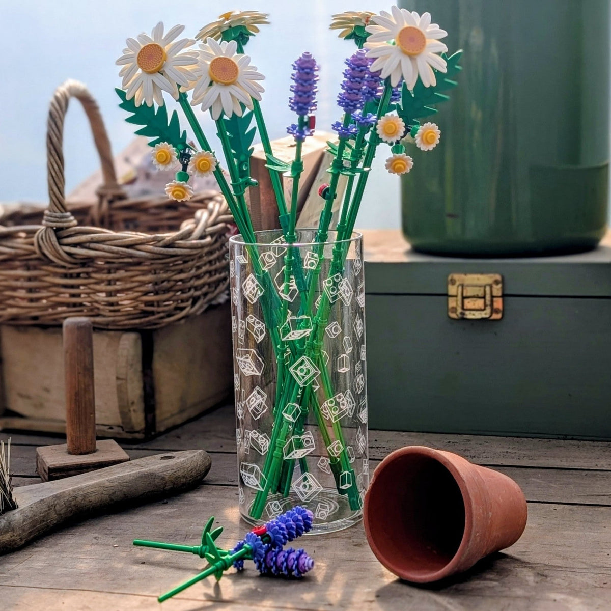 A styled still life on a wooden table with a clear glass lego vase holding artificial daisy and lavender stems, alongside a wicker basket, green box, terracotta pot, brush, and a blurred outdoor background.