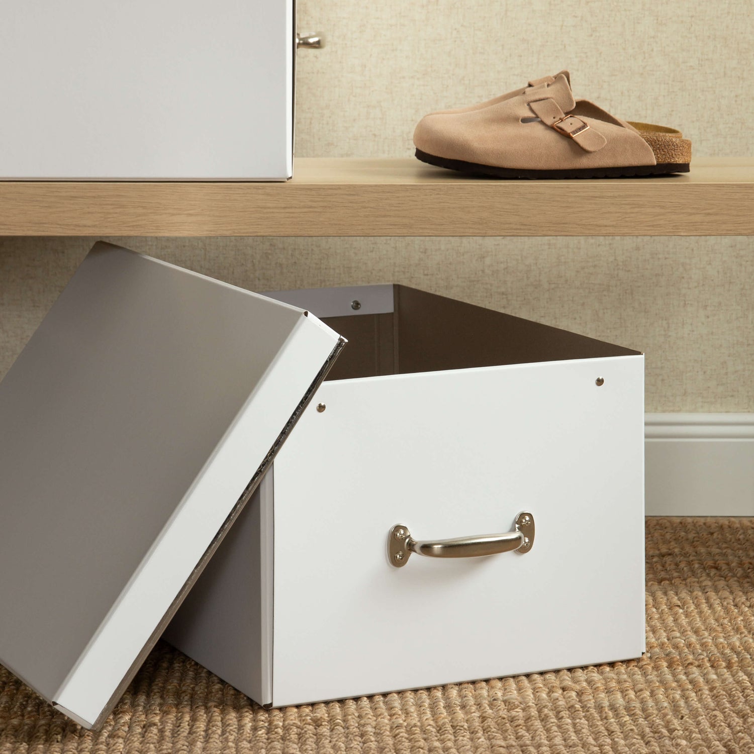 White storage box with a lid and handle on a carpeted floor, with a beige wall and wooden shelf in the background.