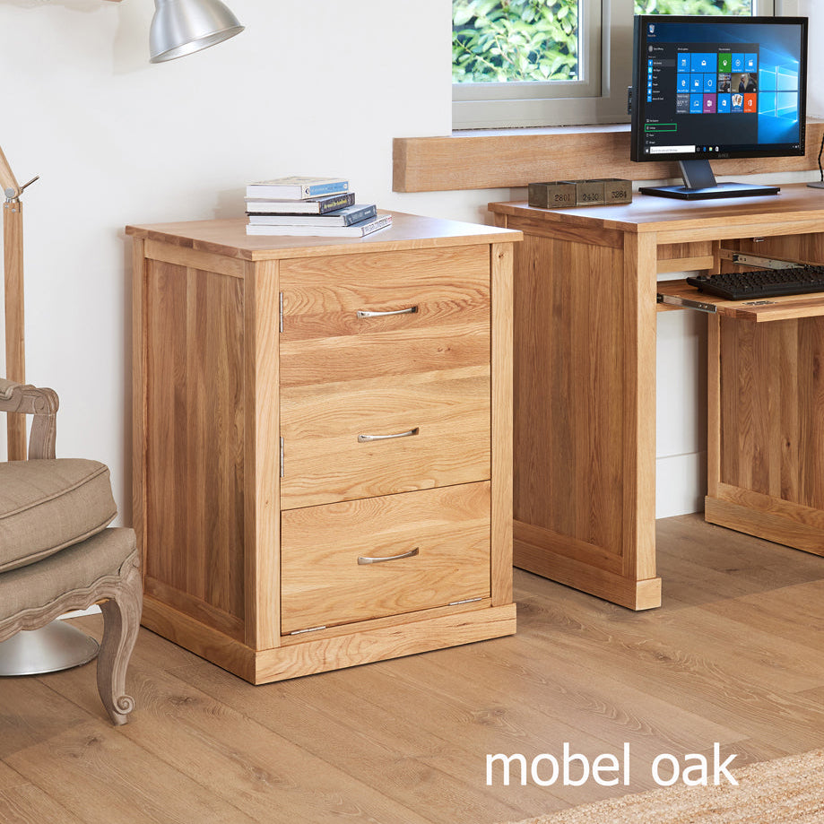 Wooden desk with computer setup and filing cabinet in a room with a window and chair.
