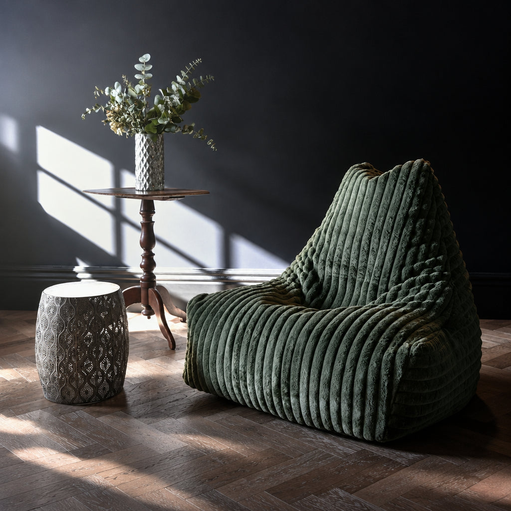 A large, plush green ribbed lounge chair with a high back, styled in a dark room with dramatic sunlight streaming across the floor, next to a small patterned side table and a vase of flowers.