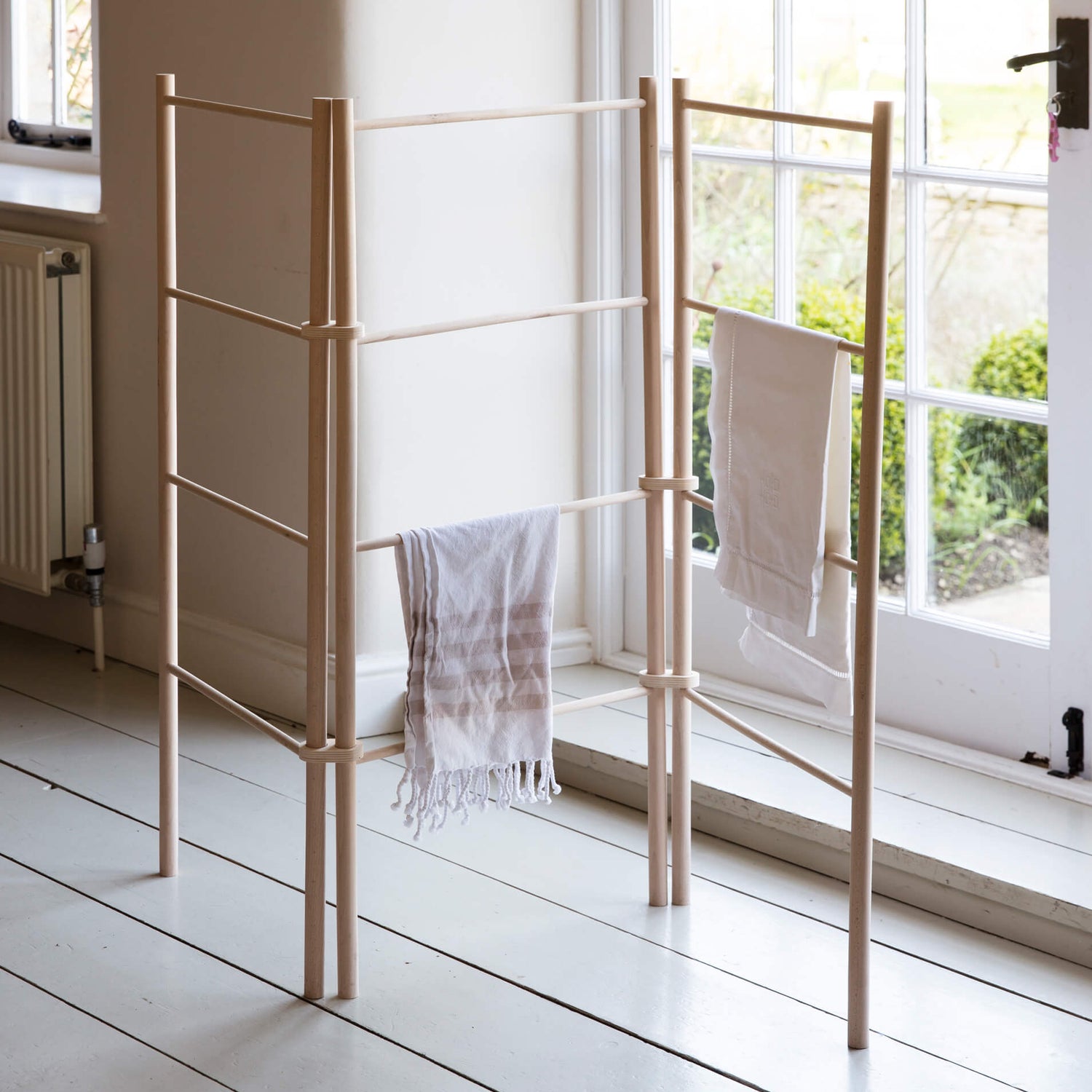 Wooden towel rack with hanging towels in a bright room with large windows.