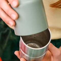 Close-up of a person's hands demonstrating a reusable travel tumbler with a removable lid. The lid is a matte sage green, while the tumbler body is brushed stainless steel silver.