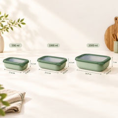 Three rectangular glass storage containers in green are shown on a light surface with printed size labels above each: 500 ml, 1000 ml, and 2000 ml. The containers have translucent lids and are photographed in a styled kitchen-like setting with plants and utensils in the background.