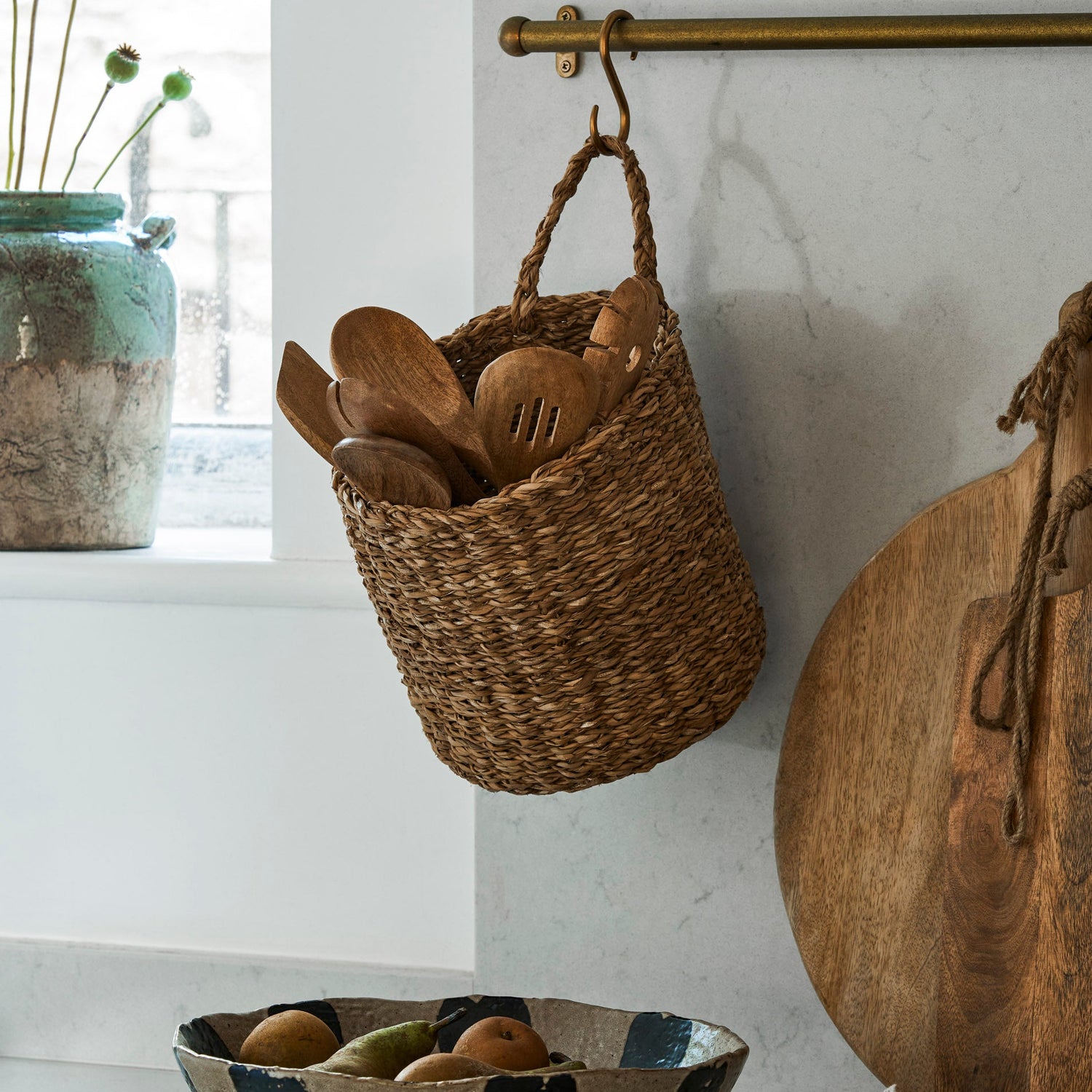 Woven basket with wooden spoons hanging on a marble wall, next to a ceramic bowl with fruits.