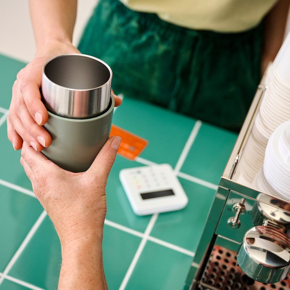 Person holding a metal cup next to a coffee machine on a tiled counter.