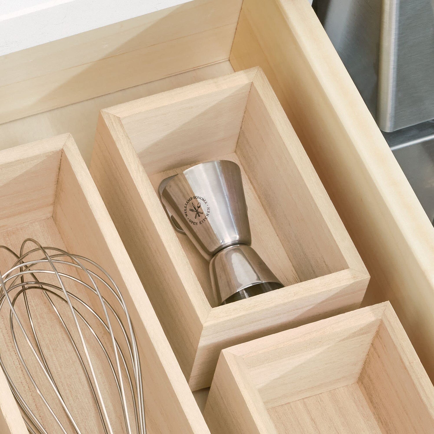 Wooden kitchen drawer organizer with a whisk and metal jigger.