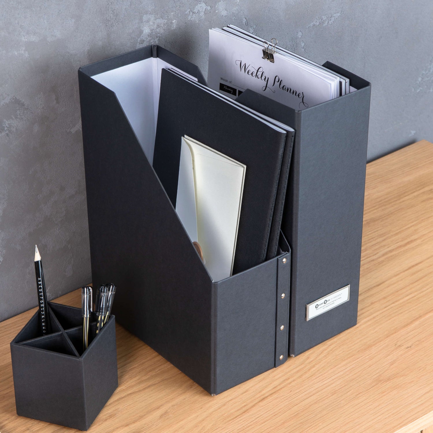 Black file folder with stationery items on a wooden surface against a gray wall.