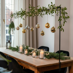 Dining table set with candles, greenery, and hanging ornaments in a room with a window and curtain.