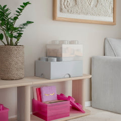 Pink gift box with gold ribbon on a wooden shelf next to a plant and textured wall art.