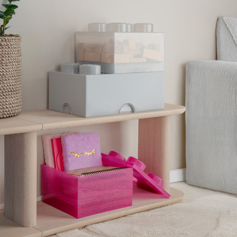 Pink gift box with gold text on a wooden shelf in a room with a plant and textured wall art.
