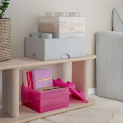 Pink gift box with gold text on a wooden shelf in a room with a plant and textured wall art.