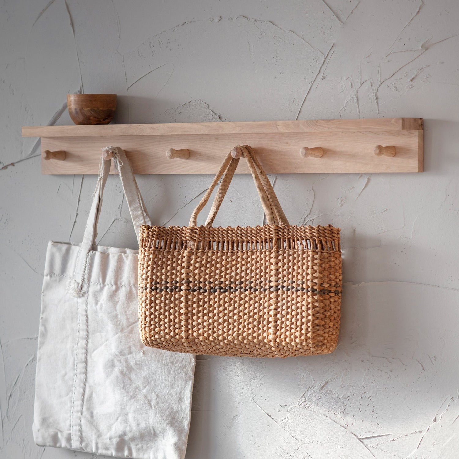 Two bags, one white and one woven, hanging on a wooden shelf against a light gray wall.