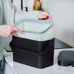 Person using a kitchen composting system with a black bin and clear bag.