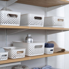 White perforated plastic storage bins arranged on light wood floating shelves against a white wall, with folded gray and blue textiles nearby.