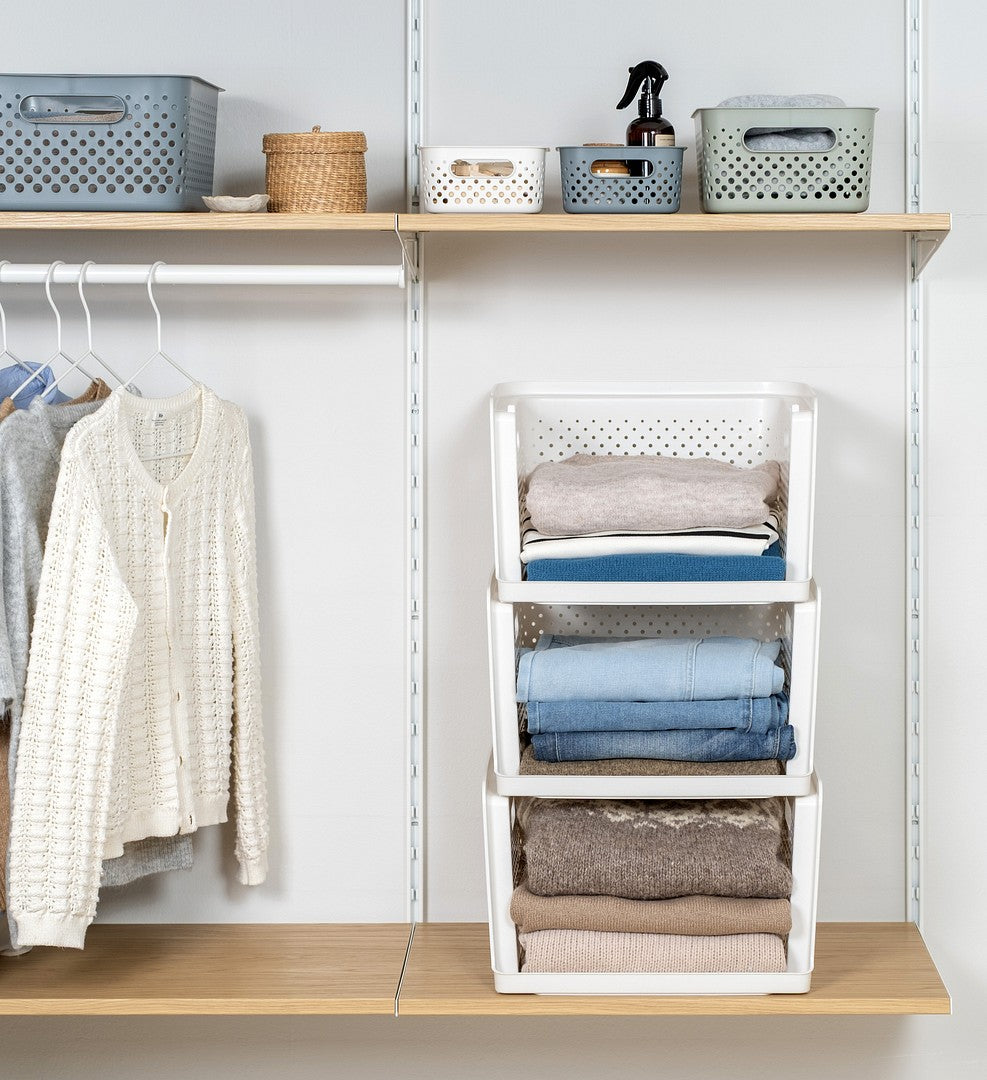 A white three-tier fold-and-fold organizer filled with folded clothing, shown in a closet setting next to hanging garments and shelves with storage bins.