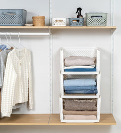 A white three-tier fold-and-fold organizer filled with folded clothing, shown in a closet setting next to hanging garments and shelves with storage bins.