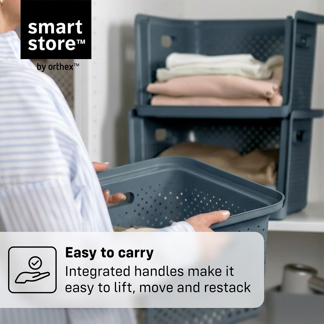 A person in a striped shirt holds a dark blue-gray plastic storage basket with an integrated handle, with a stack of folded clothes in another basket in the background. Text overlays read 'smart store by orthex' and 'Easy to carry Integrated handles make it easy to lift, move and restack'.