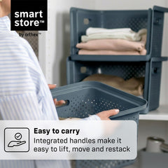 A person in a striped shirt holds a dark blue-gray plastic storage basket with an integrated handle, with a stack of folded clothes in another basket in the background. Text overlays read 'smart store by orthex' and 'Easy to carry Integrated handles make it easy to lift, move and restack'.