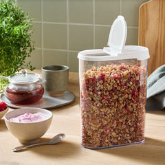 Container of granola on a wooden table with bowls of yogurt and berries, a teacup, and a plant.