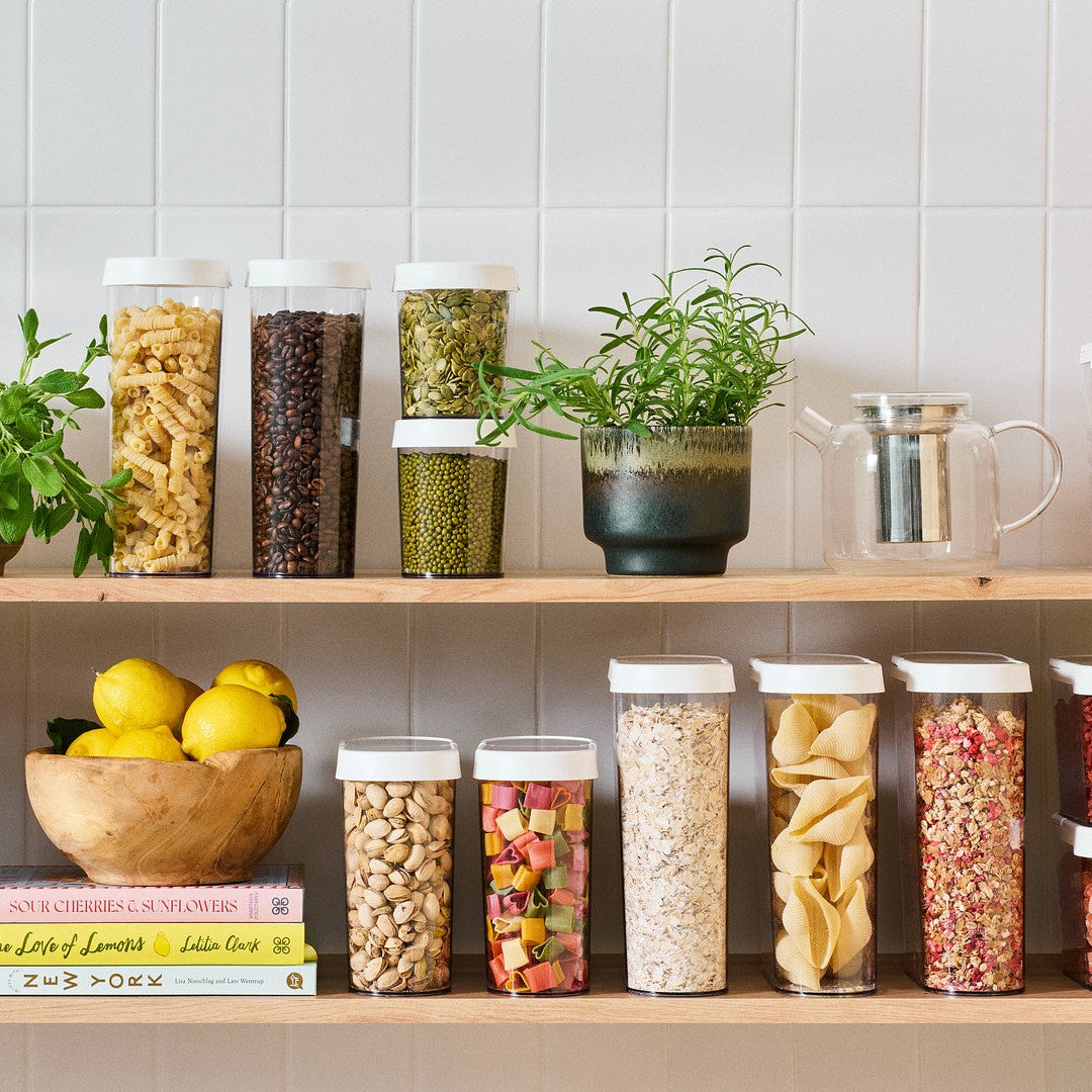 Shelves with various jars of food, plants, and books against a tiled wall.