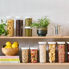 Shelves with various jars of food, plants, and books against a tiled wall.