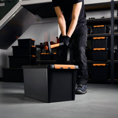 Person using a tool to open a black storage box with orange handles in a workshop setting.