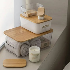 A stack of three clear plastic bathroom storage containers with bamboo lids sits on a windowsill. The containers hold rolled towels, a candle, toiletries, and a sponge brush.