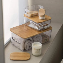 A stack of three clear plastic bathroom storage containers with bamboo lids sits on a windowsill. The containers hold rolled towels, a candle, toiletries, and a sponge brush.